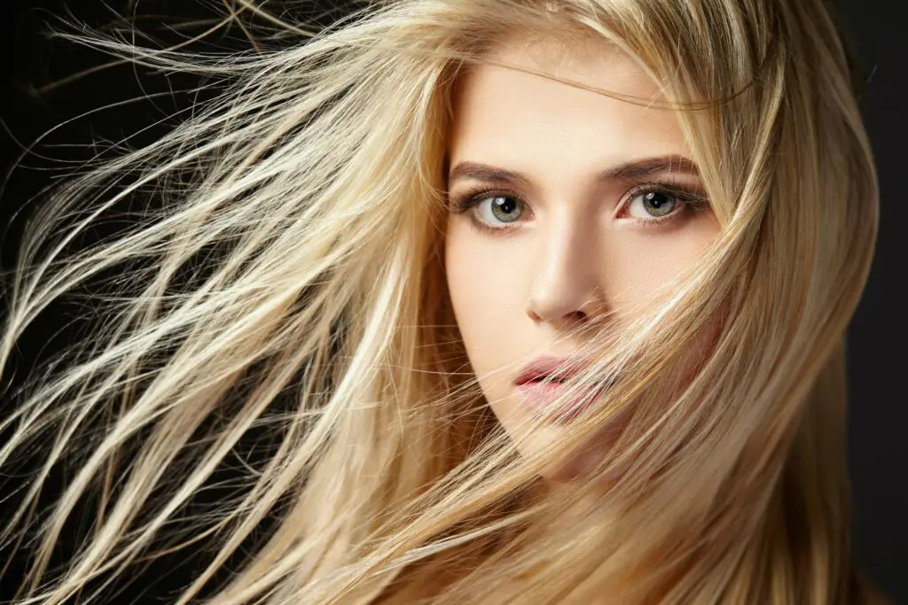 A close-up of a person with long, flowing blonde hair being blown by the wind. They have light green eyes, a calm expression, and a dark background that highlights their features. Their hair partially covers their face, creating a dynamic and captivating composition—reminiscent of an ad for premier hair salons Cary NC.