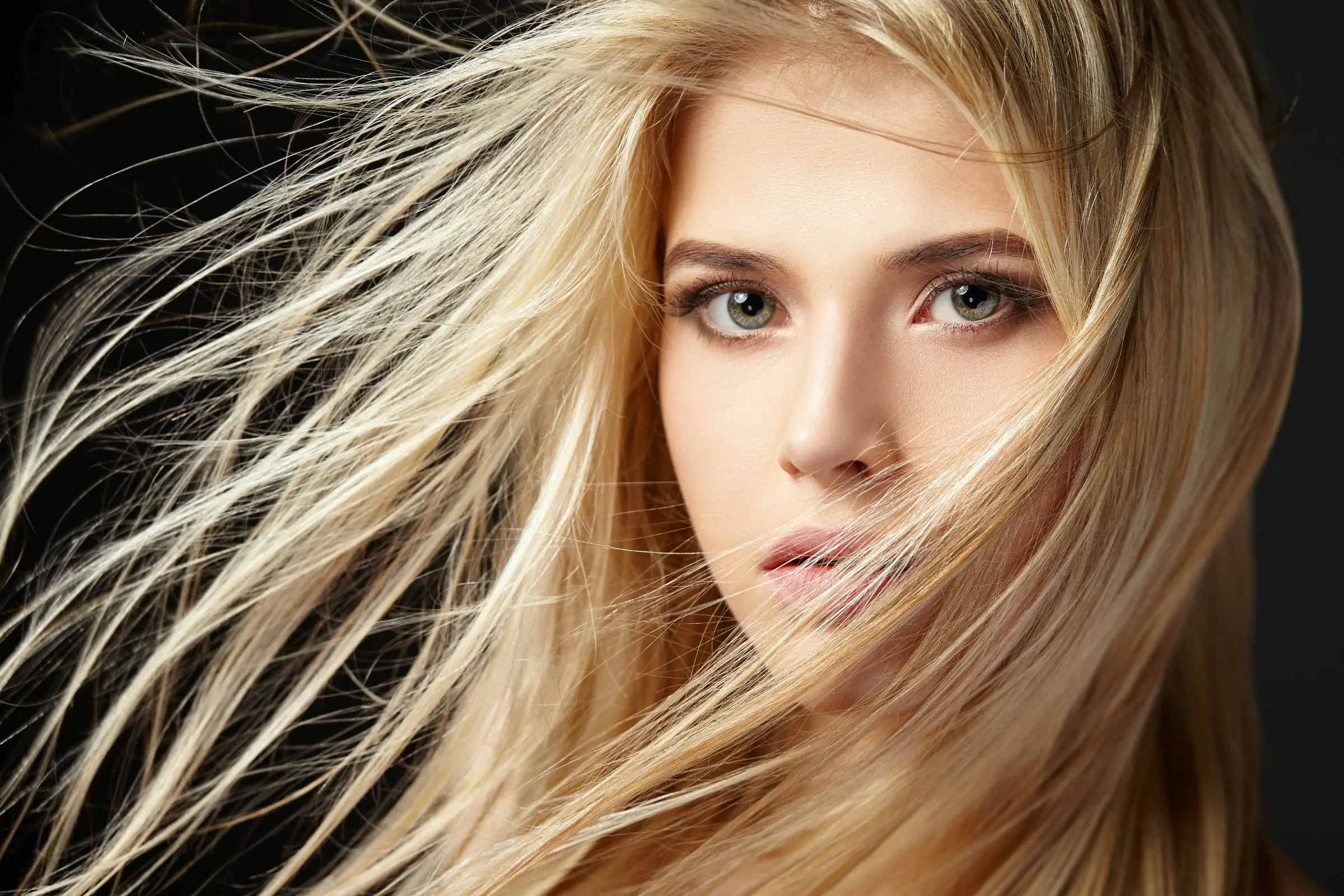 A close-up of a person with long, flowing blonde hair being blown by the wind. They have light green eyes, a calm expression, and a dark background that highlights their features. Their hair partially covers their face, creating a dynamic and captivating composition—reminiscent of an ad for premier hair salons Cary NC.