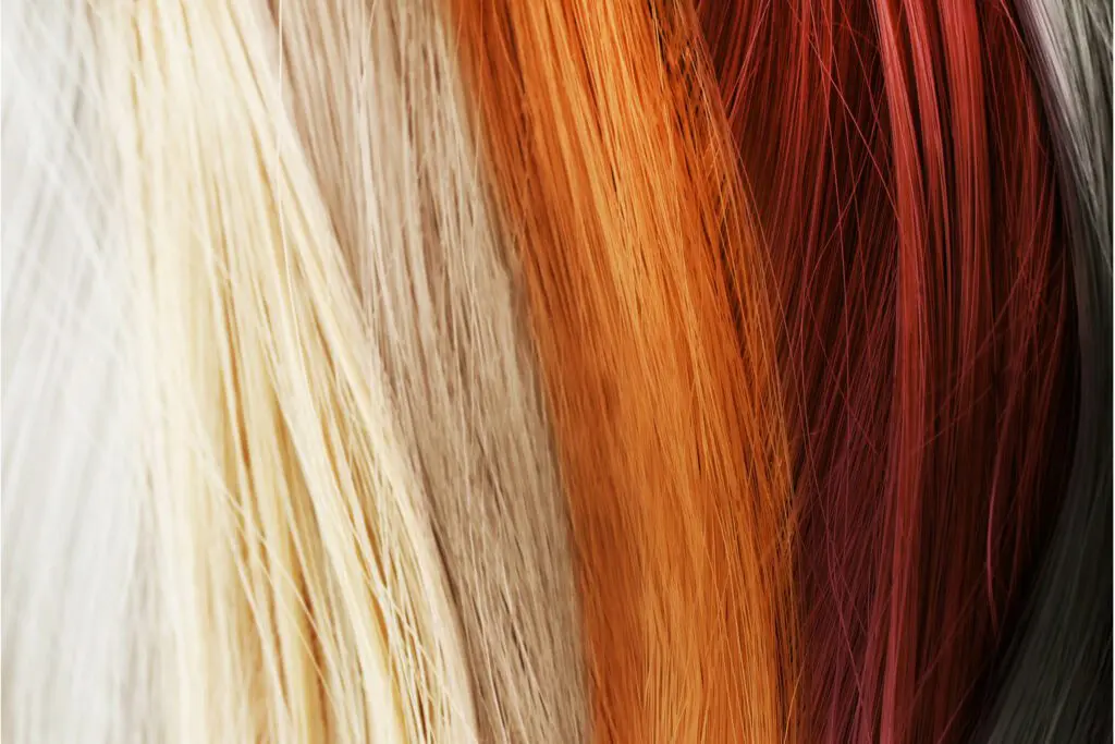 Close-up view of various strands of hair in different shades, ranging from platinum blonde to light brown, orange, deep red, and black. The strands are placed next to each other, showing a gradient of colors—an inspiring palette for your next visit to a hair salon.