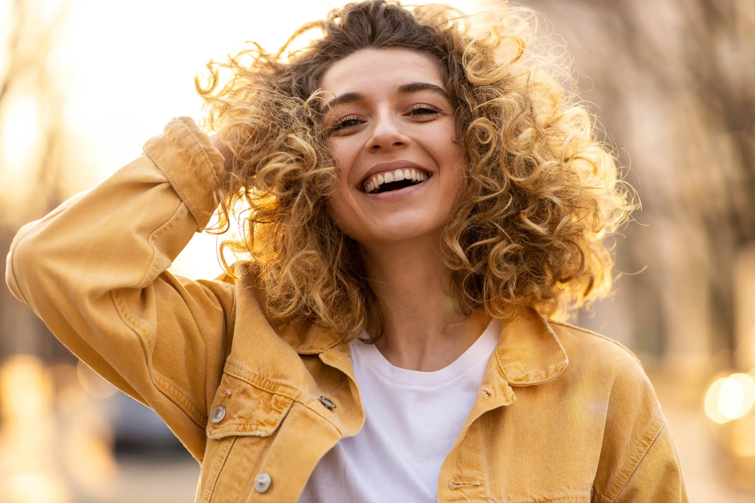 A person with curly hair is smiling and holding up their hand to their head. They are wearing a yellow jacket over a white shirt. The background is softly blurred, with warm lighting suggesting it's either sunrise or sunset—perfect for showcasing the work of the best hair salon Cary NC has to offer.
