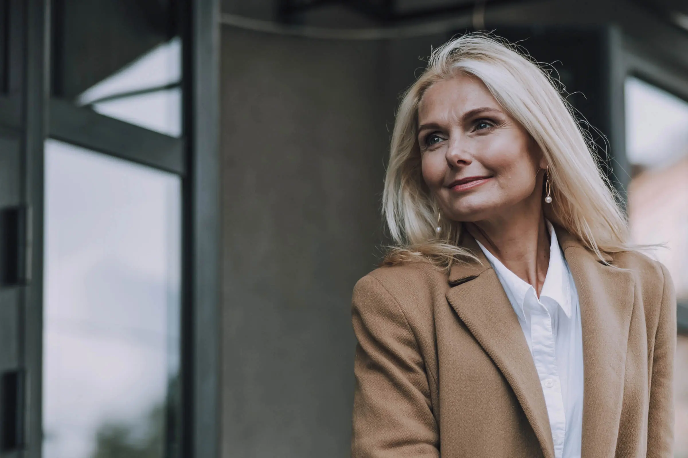 A woman with long blonde hair, styled to perfection from one of the best hair salons Cary NC offers, wears a tan coat and white blouse while standing outdoors. She smiles slightly in front of a modern building with large windows, exuding a casual yet confident expression.