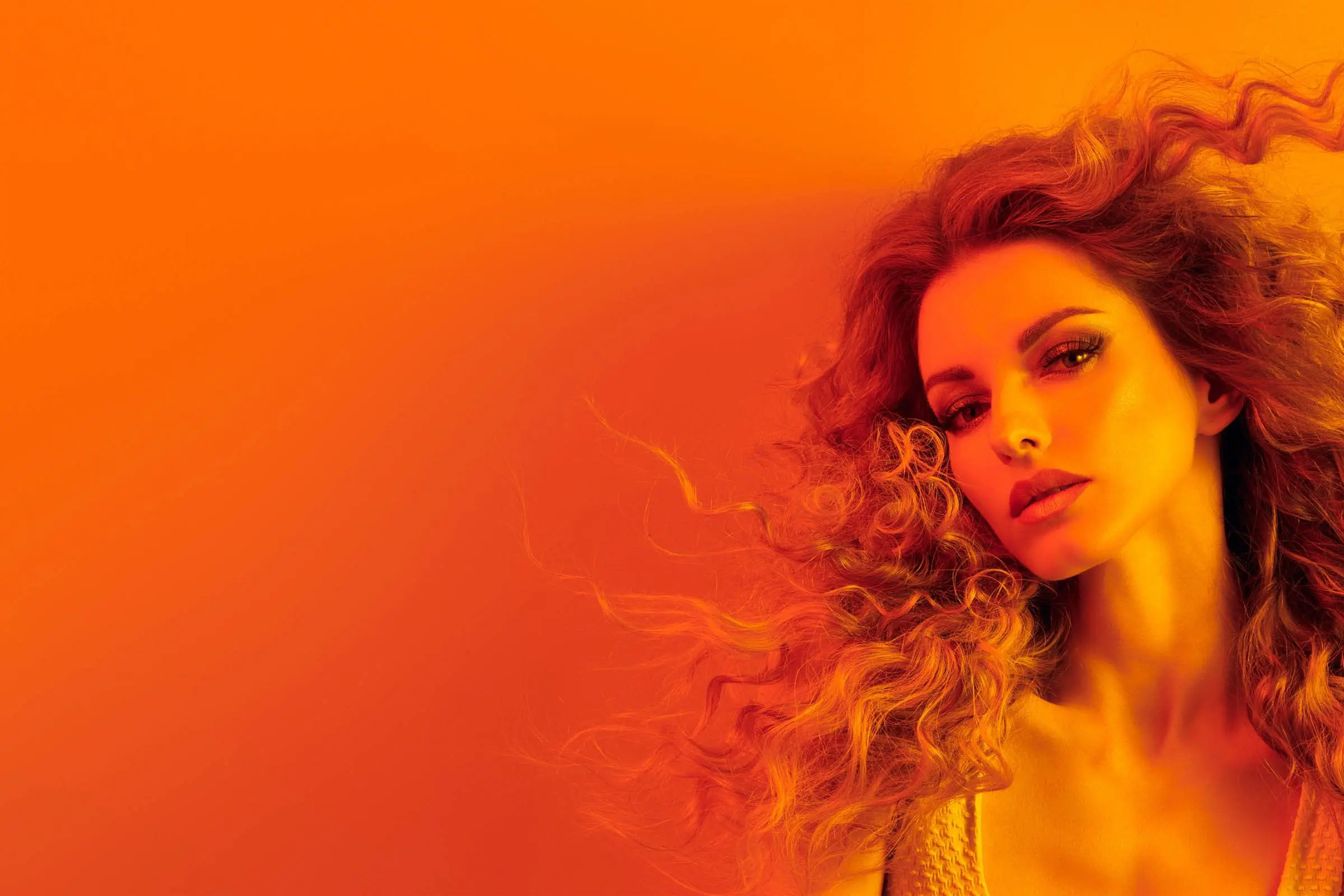 A person with voluminous, curly hair poses against a vibrant, orange gradient background. They have an intense expression and are looking slightly off to the side. The overall image is bathed in warm, orange tones, creating a dramatic and bold effect, reminiscent of trendy styles found at top hair salons near me.