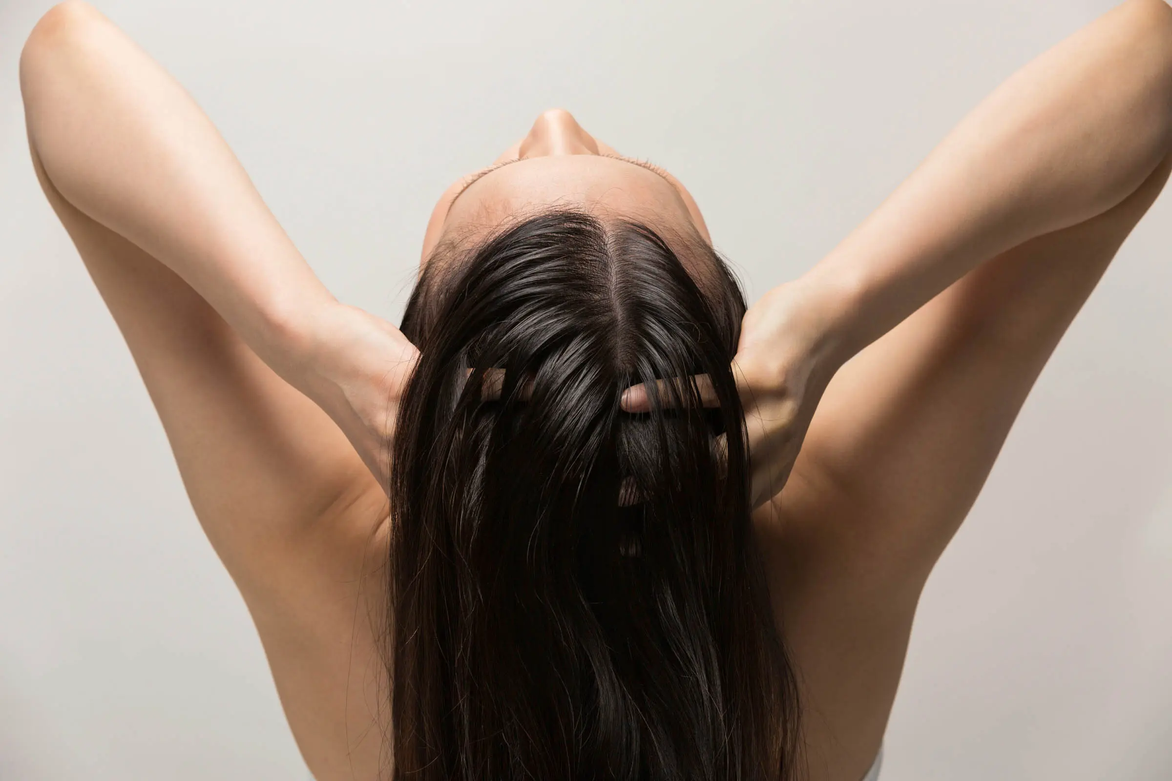 A person with long dark hair is seen from behind, with their head tilted back and their hands massaging their scalp. Their elbows are pointed outward, and the background is plain and light-colored—a serene moment that could easily be a scene from a Sunday salon experience at the best hair salon Cary NC offers.