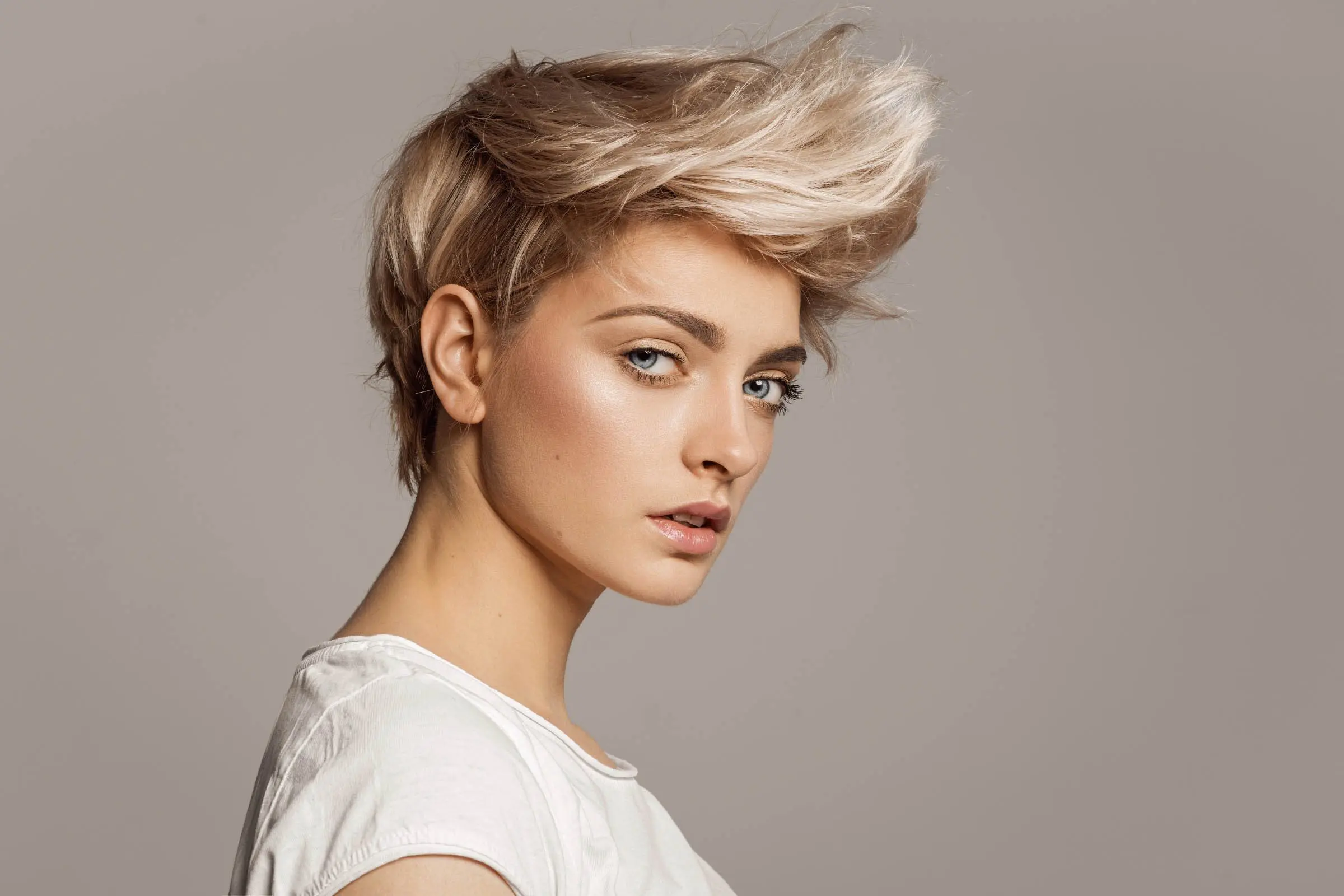 A person with short, platinum-blonde hair styled in an edgy, voluminous look gazes confidently at the camera. They are wearing a white shirt and are set against a plain, light grey background. Their makeup is subtle, emphasizing their eyes and cheekbones; it's clear they've visited the best hair salon in Cary NC.