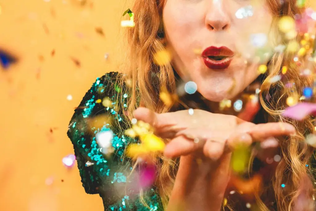 A person with long, curly hair is blowing a kiss, causing a burst of colorful confetti to scatter. The individual, possibly styled by one of the best hair salons in Raleigh, NC, is wearing a glittery green outfit against a warm orange backdrop. The image conveys a celebratory and festive atmosphere.