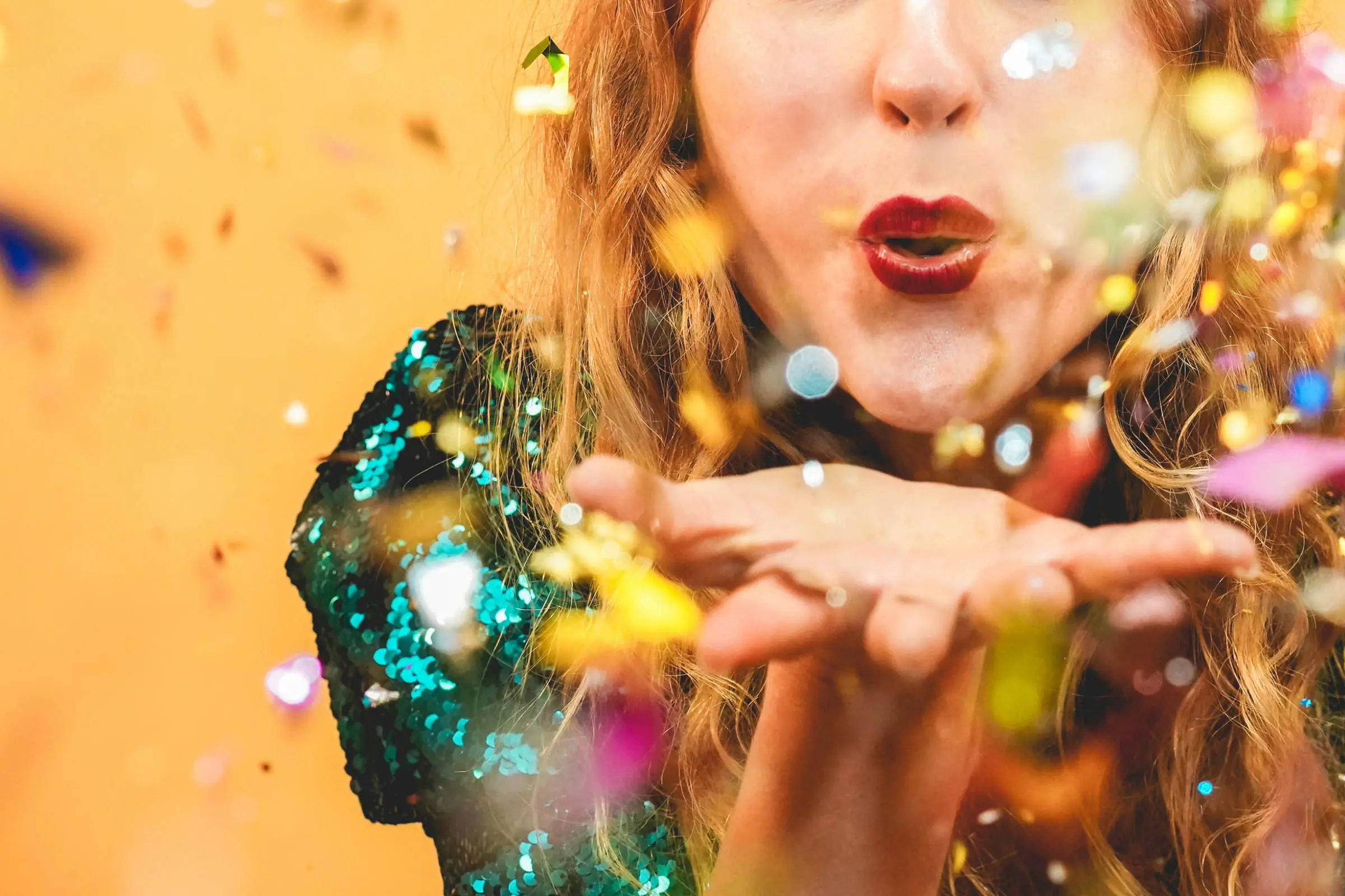 A person with long, curly hair is blowing a kiss, causing a burst of colorful confetti to scatter. The individual, possibly styled by one of the best hair salons in Cary, NC, is wearing a glittery green outfit against a warm orange backdrop. The image conveys a celebratory and festive atmosphere.