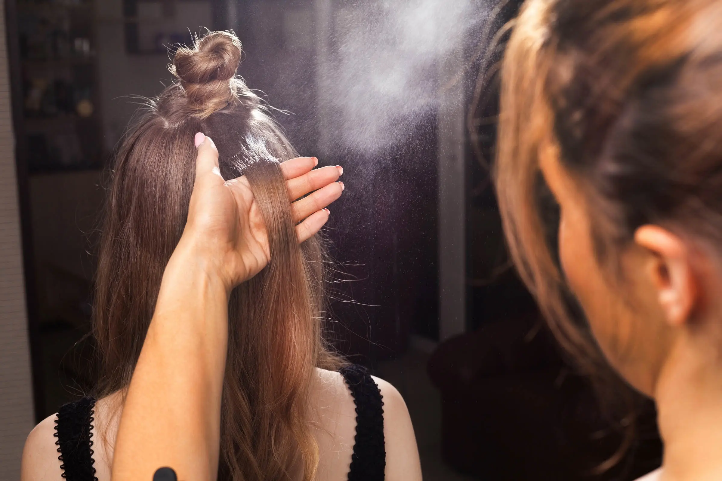 A person at the best hair salon in Cary, NC, sprays hairspray on the back of a woman’s head while styling her hair. The woman’s hair is partially tied in a top knot with a section loose, and the stylist is holding a strand of hair, preparing to perfect it.