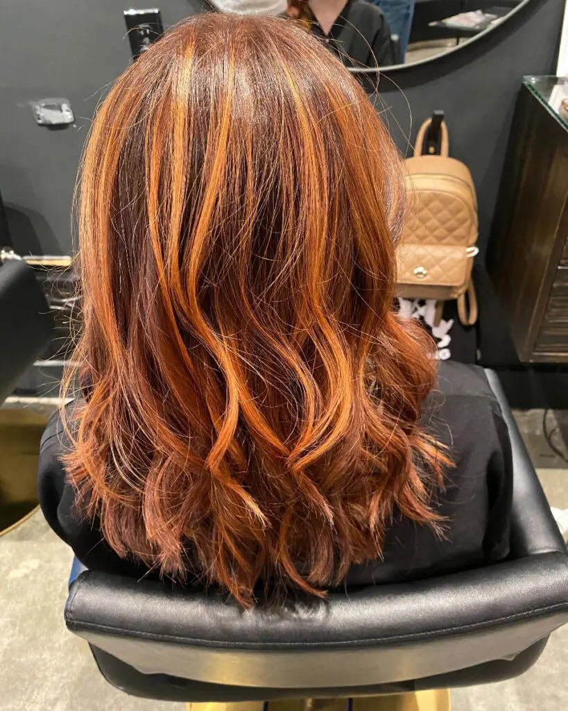 Back view of a woman sitting in a salon chair showcasing medium-length wavy hair with high-contrast fiery orange and copper ribbon highlights over a deep auburn base, custom colored by Lisa Edwards at Sunday Salon in Raleigh, NC.