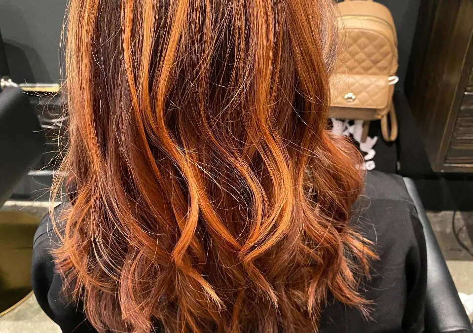 Back view of a woman sitting in a salon chair showcasing medium-length wavy hair with high-contrast fiery orange and copper ribbon highlights over a deep auburn base, custom colored by Lisa Edwards at Sunday Salon in Raleigh, NC.