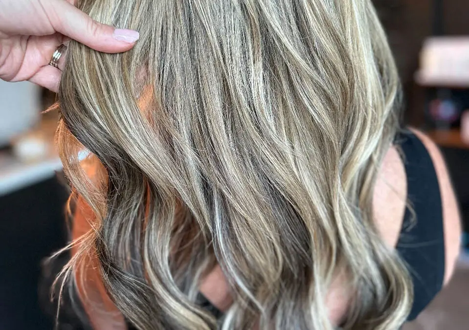 A stylist's hand showcasing long, wavy hair featuring high-contrast vanilla blonde ribbon highlights over a dimensional bronde base, custom colored by Lizzie Post at Sunday Salon in Raleigh, NC.