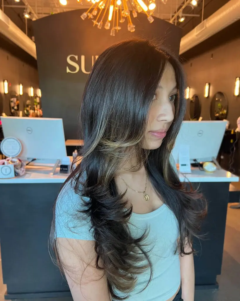 Woman showcasing long, dark brunette hair styled with voluminous sweeping layers and a high-contrast blonde face-framing money piece, custom colored by Lizzie Post at Sunday Salon in Raleigh, NC.