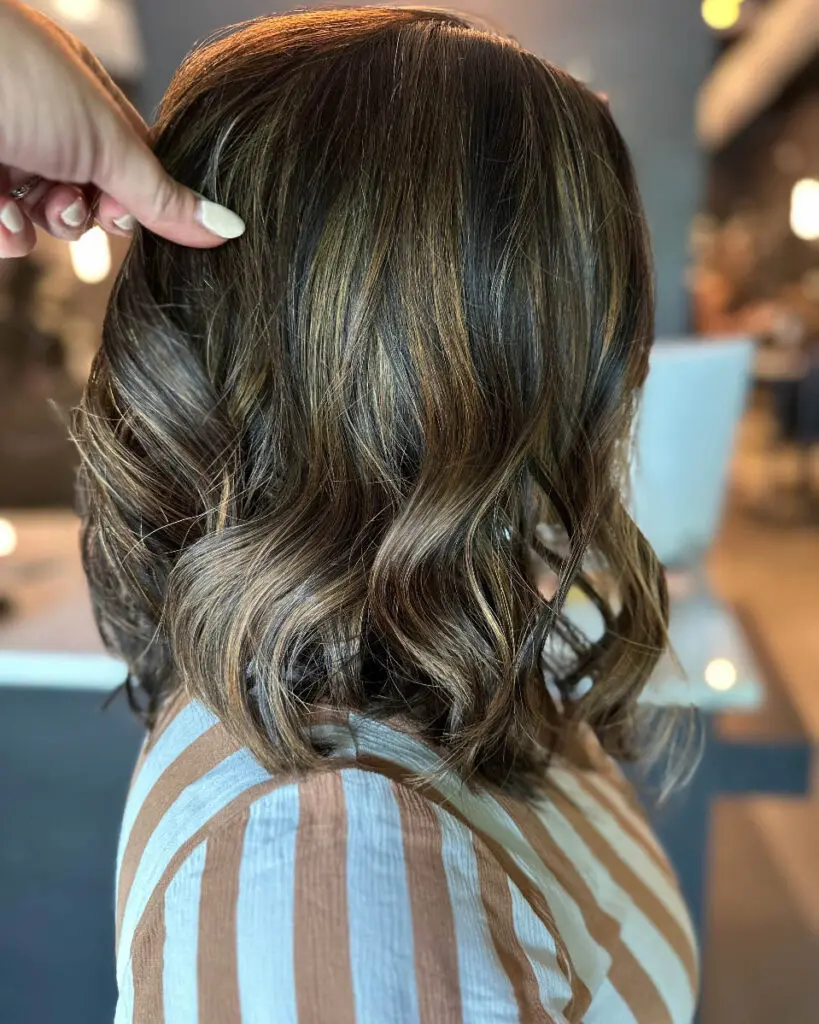 A stylist's hand showcasing a textured, wavy lob haircut featuring a deep brunette base with high-contrast golden caramel balayage highlights, custom colored by Lizzie Post at Sunday Salon in Raleigh, NC.