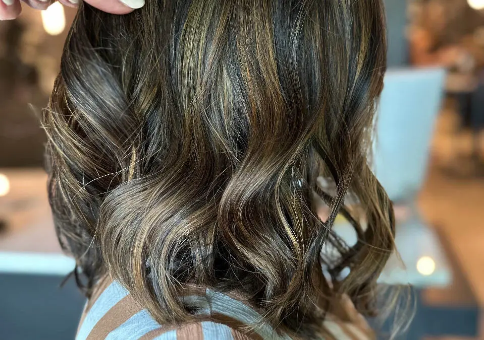 A stylist's hand showcasing a textured, wavy lob haircut featuring a deep brunette base with high-contrast golden caramel balayage highlights, custom colored by Lizzie Post at Sunday Salon in Raleigh, NC.