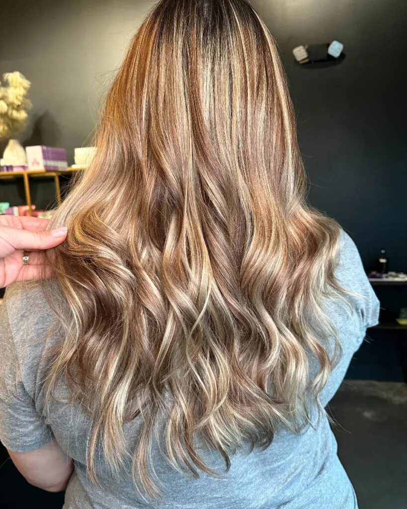 Back view of a woman with ultra-long, thick hair showcasing a dimensional warm caramel and honey bronde balayage styled in loose cascading waves, custom colored by Lizzie Post at Sunday Salon in Raleigh, NC.
