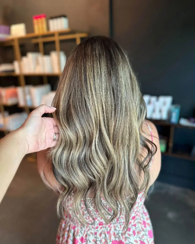 High-Contrast Creamy Blonde Balayage with Long Cascading Waves A stylist's hand showcasing long, wavy hair featuring high-contrast creamy blonde ribbon highlights over a deep brunette base, custom colored by Lizzie Post at Sunday Salon in Raleigh, NC.