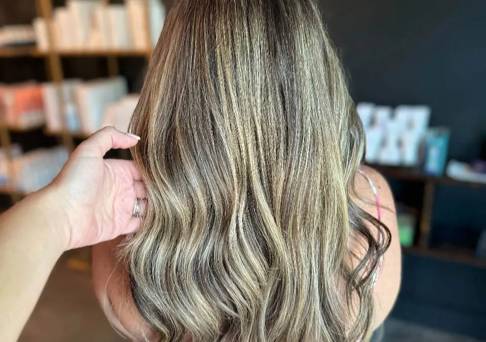 A stylist's hand showcasing long, wavy hair featuring high-contrast creamy blonde ribbon highlights over a deep brunette base, custom colored by Lizzie Post at Sunday Salon in Raleigh, NC.