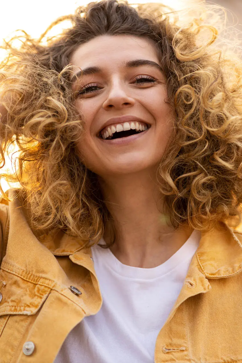A young woman with curly blonde hair smiles brightly at the camera. She is wearing a white T-shirt underneath a yellow denim jacket. The background is softly lit, contributing to a warm and joyful atmosphere.