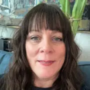 A woman with long, wavy brown hair and straight bangs is smiling at the camera. She is seated indoors on a blue sofa, with a painting and green plant visible in the background. If you're looking for similar styles, check out hair salons in Cary, NC for expert care.