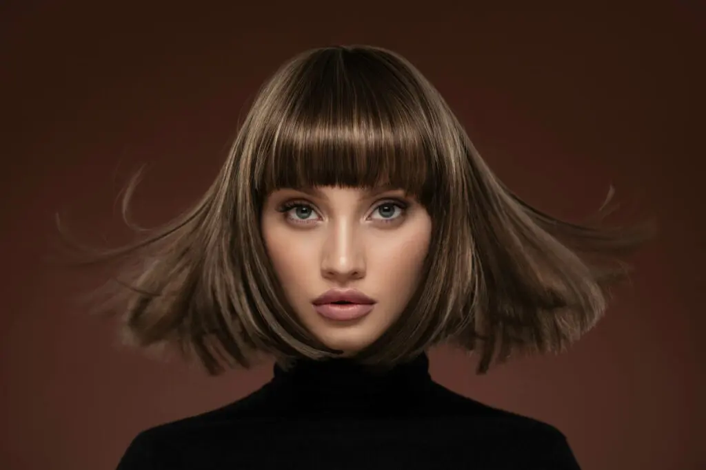 A woman with straight, shoulder-length brown hair and bangs stares directly at the camera. Her hair fans out smoothly, creating motion. She is wearing a black turtleneck against a solid brown background. Her expression is neutral and focused – perfectly capturing the essence of elegance you’d find at top hair salons in Cary, NC.