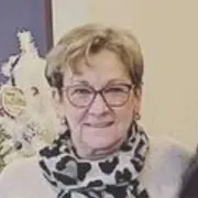 A person with light brown short hair and wearing glasses is smiling. They are dressed in a light-colored sweater and a patterned scarf, likely styled after visiting the best hair salon in Cary, NC. The background shows a decorative object that might be a flower arrangement.