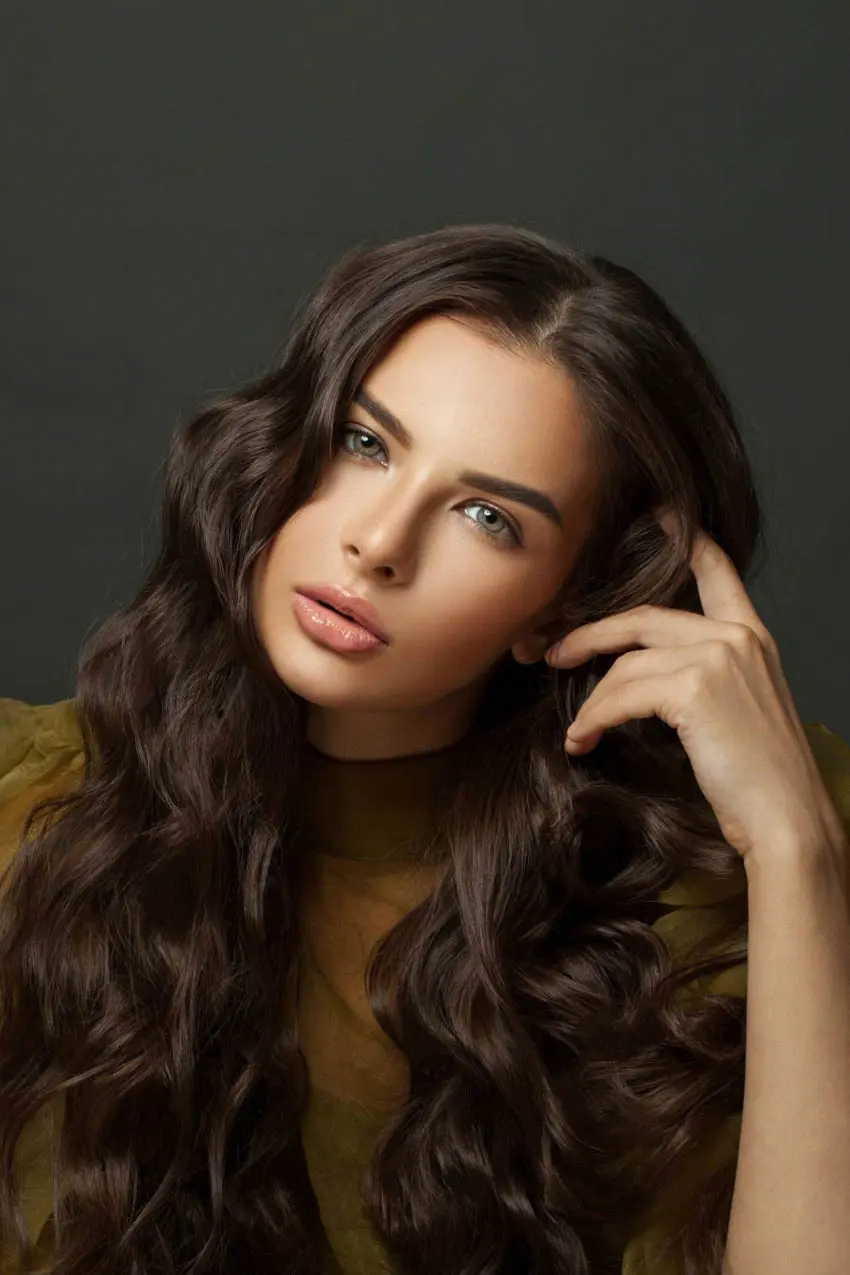 A woman with long, wavy brown hair poses against a dark background. She has green eyes and a serious expression, with her right hand touching her hair and her left hand resting near her face. She wears a see-through olive green top.