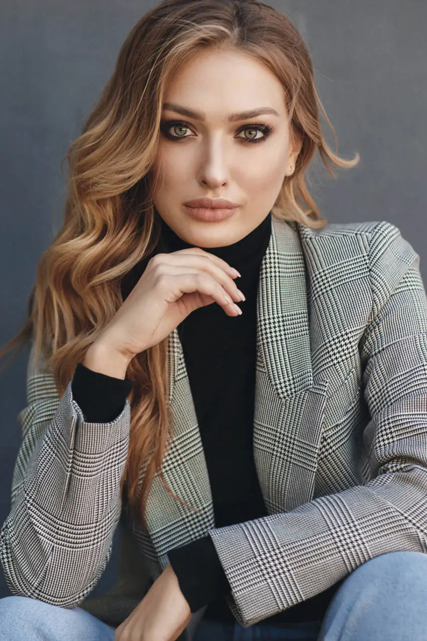 A woman with long, wavy hair is seated against a plain background. She is wearing a black turtleneck and a plaid blazer. Her hand is resting near her chin, and she is looking directly at the camera with a serious expression.