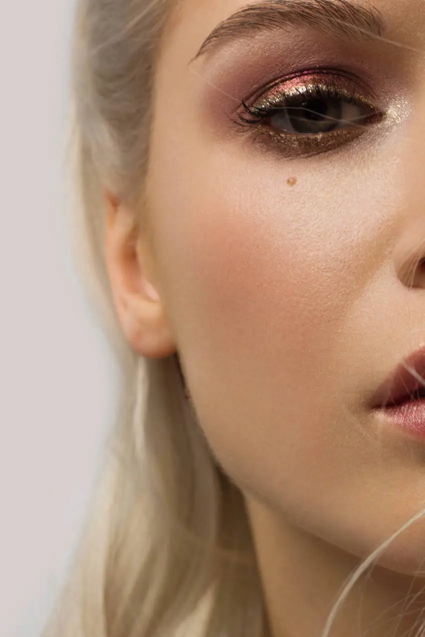 Close-up of the right side of a person's face showcasing detailed makeup. The eye features shimmering gold and pink eyeshadow with thick eyeliner. The skin appears smooth with highlighted cheekbones, and there is a beauty mark below the eye. Pale hair is visible.