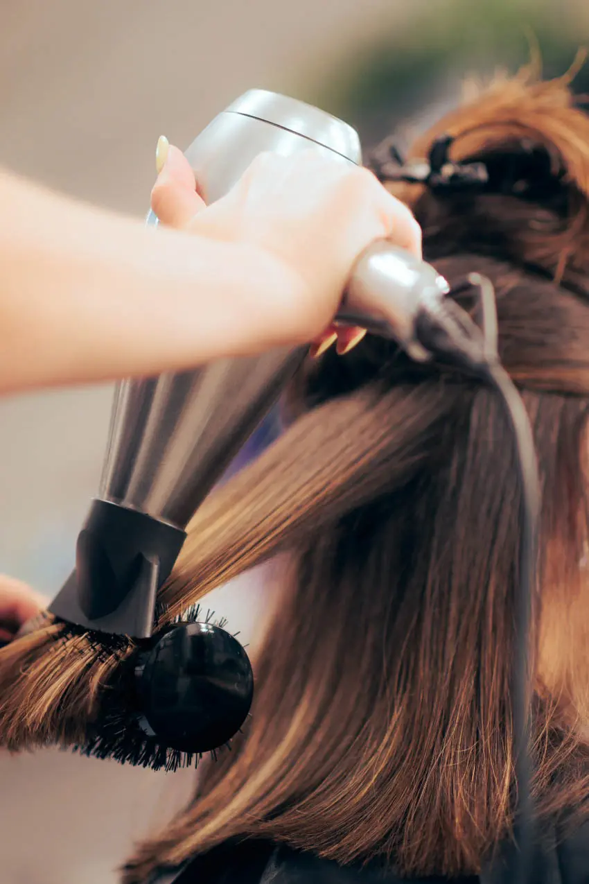 At a top hair salon in Cary, NC, a skilled hairdresser uses a blow dryer and a round brush to style a client's hair. The client's hair is sectioned and partially pinned up, with the focus on the meticulous action of blow-drying and brushing.