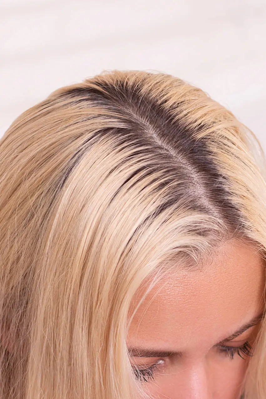 Close-up of the top of a person's head showing dark roots with blond hair. The hair is parted in the middle, highlighting the contrast between the natural dark roots and the dyed blond hair. The person's face is partially visible at the bottom of the image.
