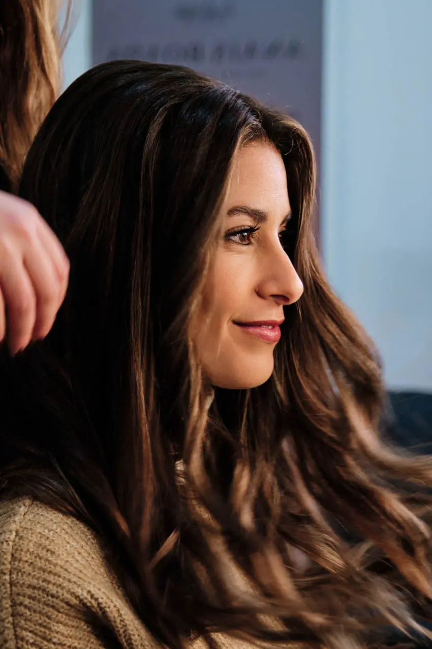 A woman with long, wavy brown hair and wearing a beige sweater is sitting in a hair salon. She's smiling slightly and looking to her right. A stylist's hand is visible, adjusting her hair. The background is blurred.