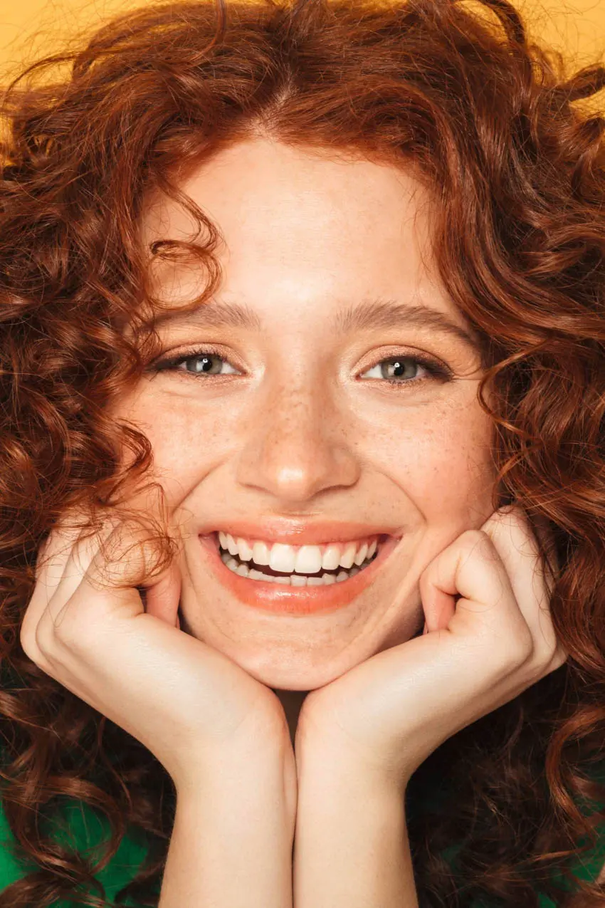 A person with curly red hair, light skin, and freckles smiles broadly, resting their chin on their hands. They are looking directly at the camera. The background is a warm, solid color.