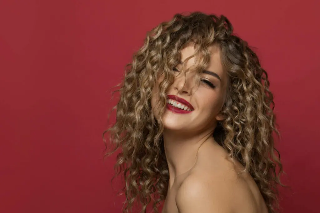 A woman with long, curly hair smiles brightly against a red background. She is showing her teeth and looking slightly off to the side, giving a joyful and lively impression—like she just left one of the top hair salons in Cary, NC.