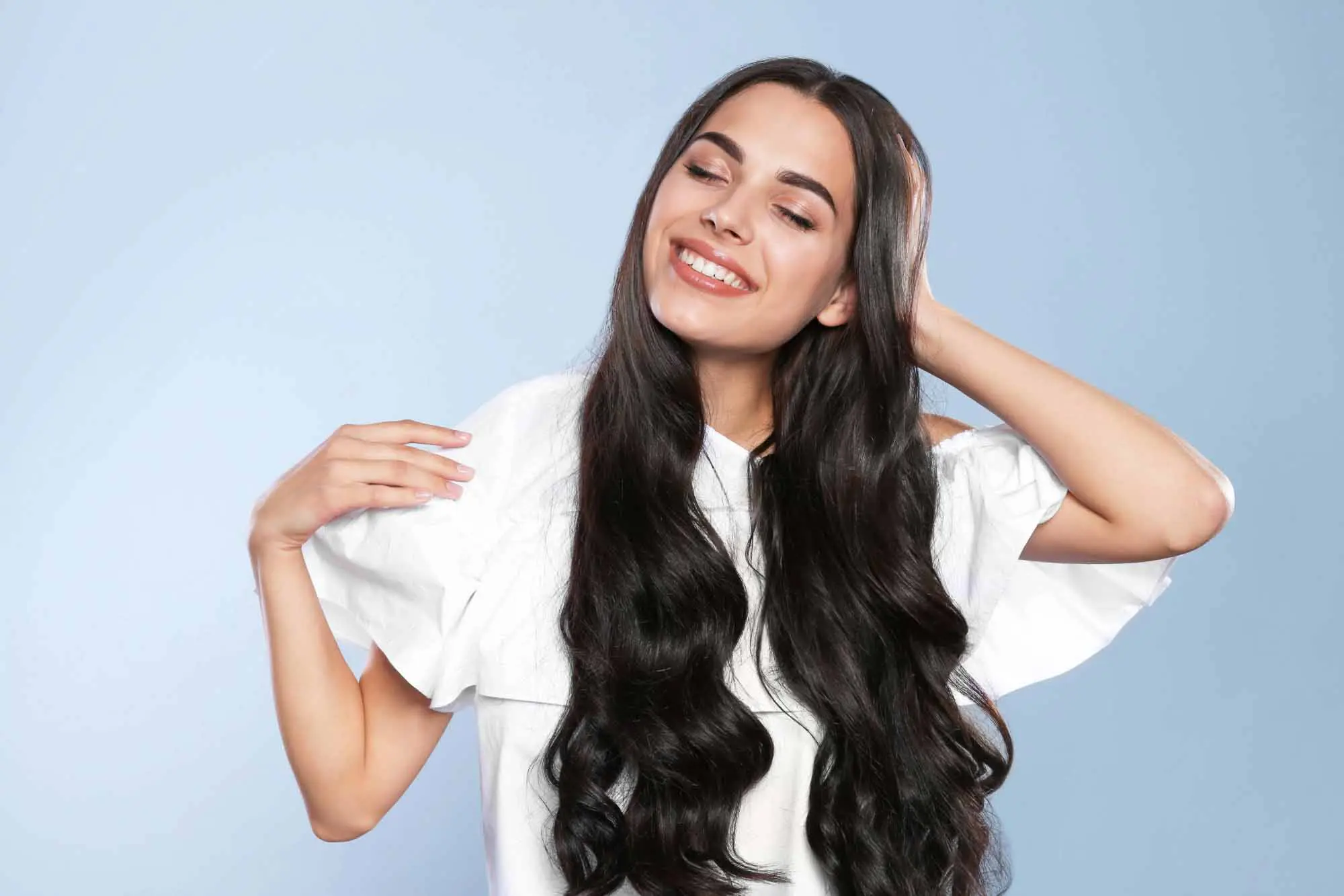 A woman with long, wavy dark hair smiles contentedly while looking slightly to the side. She is wearing a white shirt and posing with one hand touching her hair and the other resting on her shoulder against a light blue background, perfectly showcasing the results from a top hair salon near me.