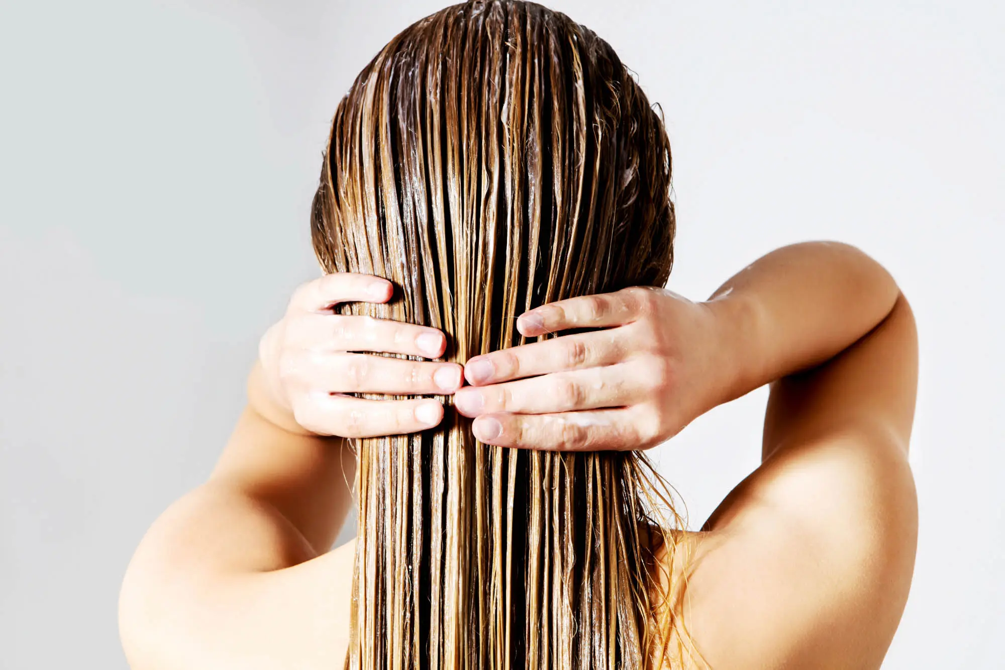 A person with wet, long, light brown hair stands facing away, with their hands placed on the back of their head. The hair is sleek and straight, as if they just stepped out of the best hair salon Cary NC has to offer. The background is a plain light gray.