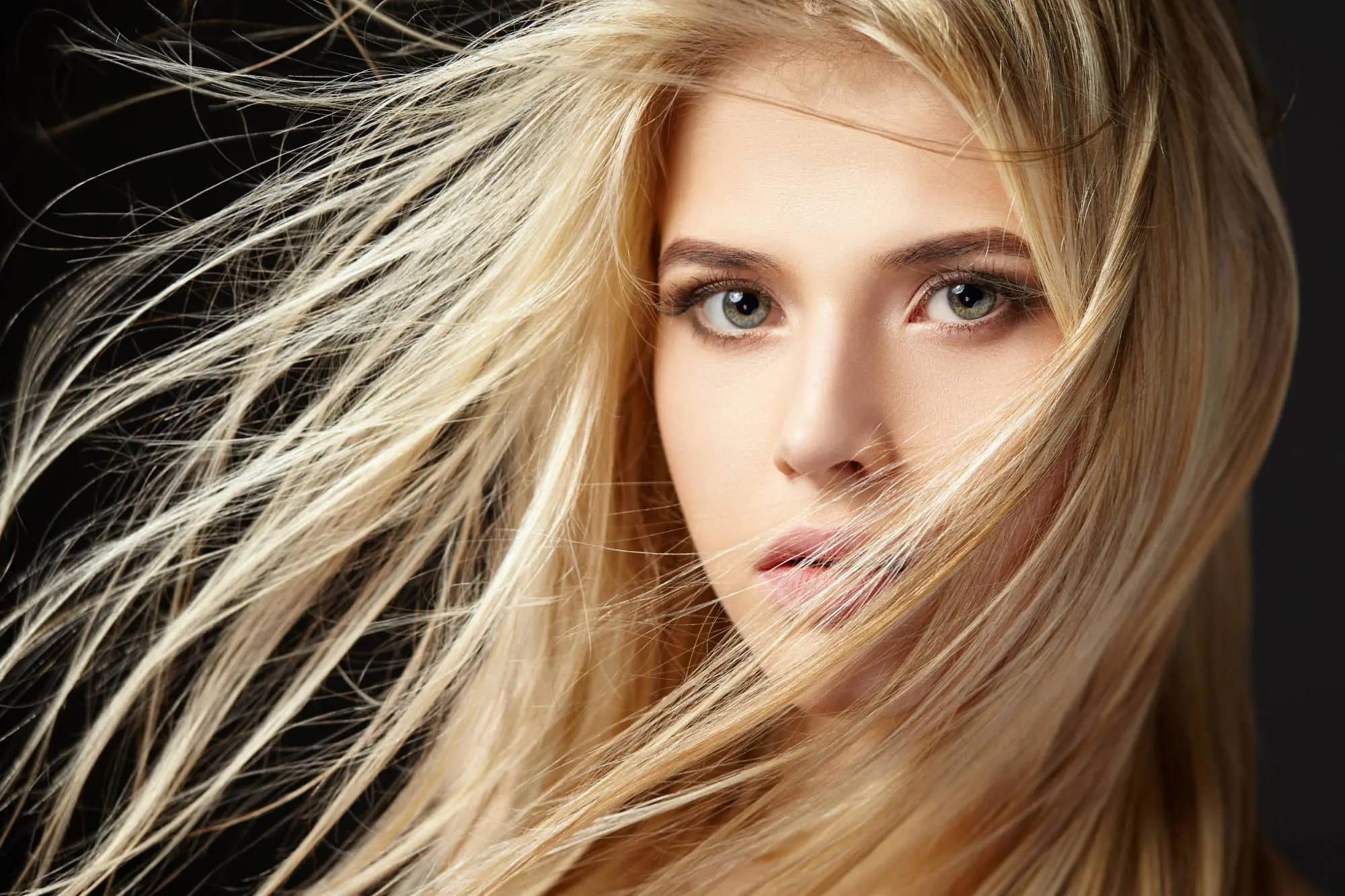 Close-up portrait of a young woman with long, flowing blonde hair subtly blowing across her face. She has green eyes and is looking directly into the camera against a dark background—an image that could grace any top hair salon's portfolio.