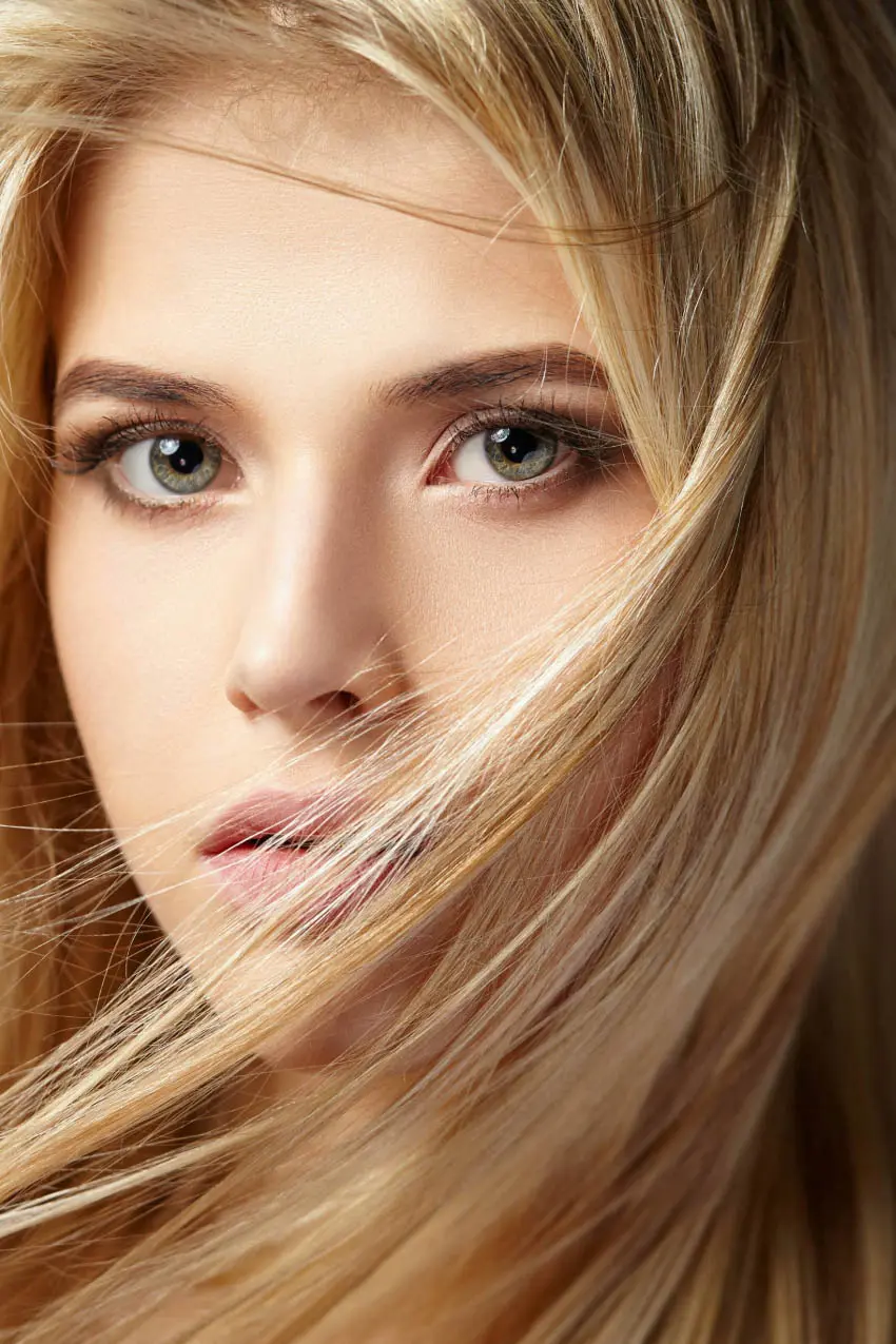 Close-up portrait of a person with long, blonde hair and striking, light-colored eyes. The hair partially covers their face, and they have a neutral expression with slightly parted lips. The background is dark, emphasizing their facial features—a look you'd expect from the best hair salon Cary NC offers.