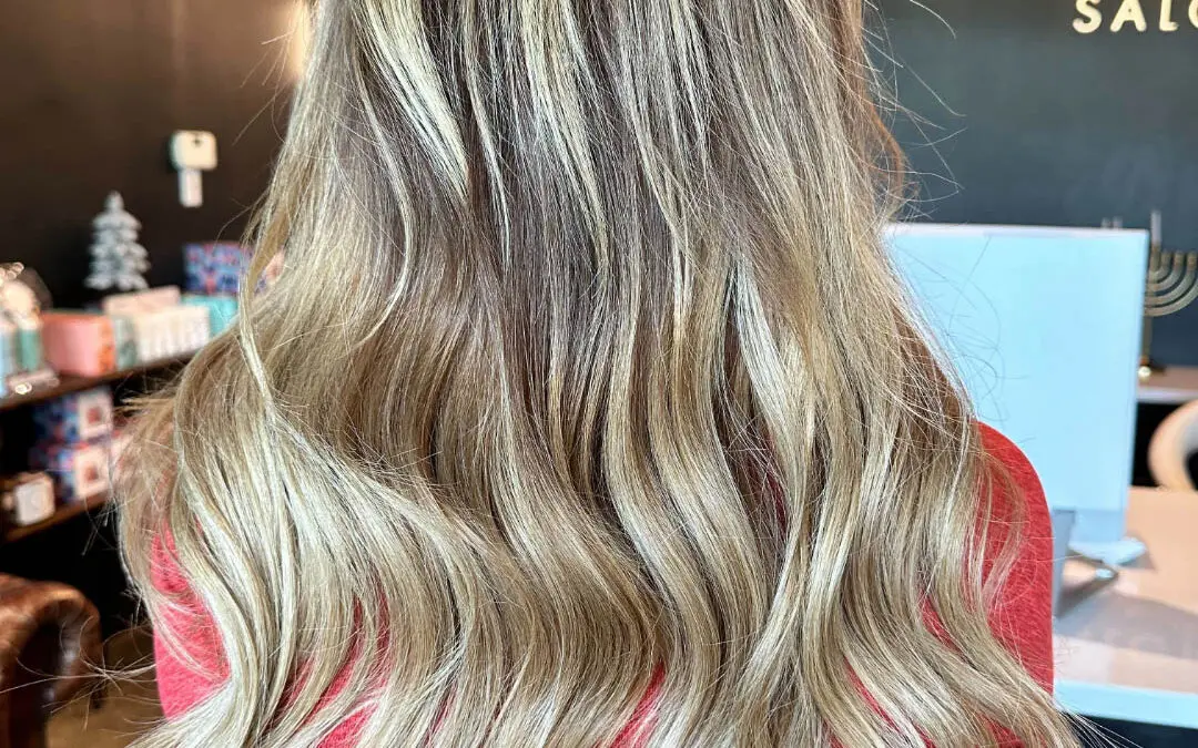 Back view of a woman showcasing ultra-long, seamlessly blended hair extensions featuring a dimensional blonde balayage and soft cascading waves, installed and custom colored by Lizzie Post at Sunday Salon in Raleigh, NC.