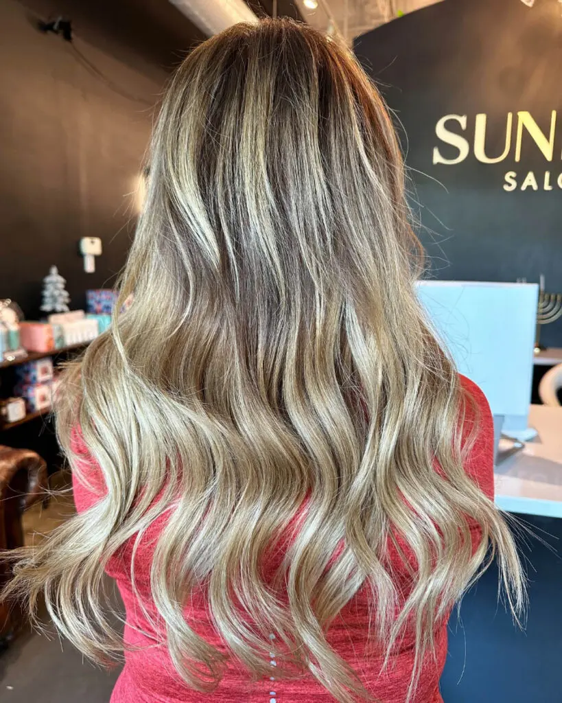 Back view of a woman showcasing ultra-long, seamlessly blended hair extensions featuring a dimensional blonde balayage and soft cascading waves, installed and custom colored by Lizzie Post at Sunday Salon in Raleigh, NC.