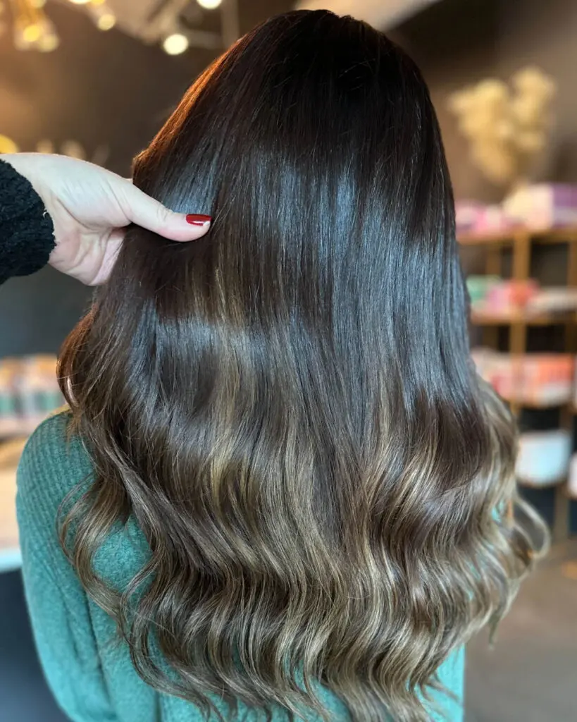 A stylist's hand showcasing ultra-thick, long dark brunette hair extensions seamlessly blended with a subtle bronde balayage, custom matched and installed by Lizzie Post at Sunday Salon in Raleigh, NC.