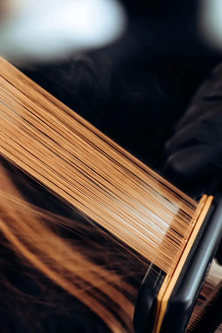 Close-up shot of a hair straightener being used on a section of long, light brown hair. The hair is being pulled taut as the flat iron clamps down, visibly straightening the strands. The background is blurred, highlighting the focus on the hair and straightener.