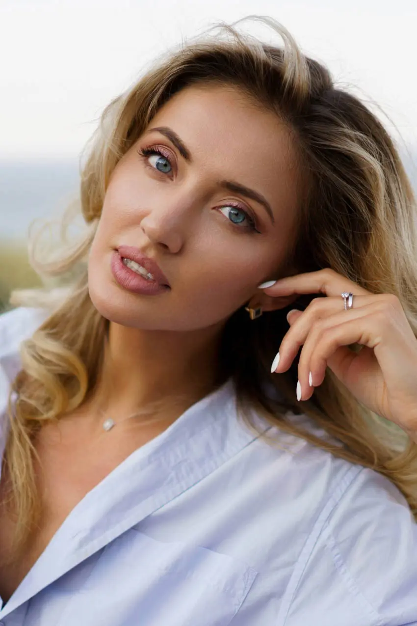 A person with long, wavy blonde hair and light blue eyes gazes towards the camera, resting their chin on their hand. They are wearing a light blue button-up shirt, and their nails are polished white. Their expression is neutral, and they are accessorized with rings and a necklace.