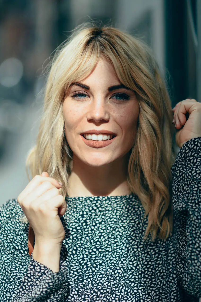 A smiling woman with blonde hair, wearing a patterned black and white top, stands outdoors. She has her left hand raised near her face and her right hand adjusting her hair, reflecting the vibrant style you can find at the best hair salons in Cary, NC. The background is beautifully blurred.
