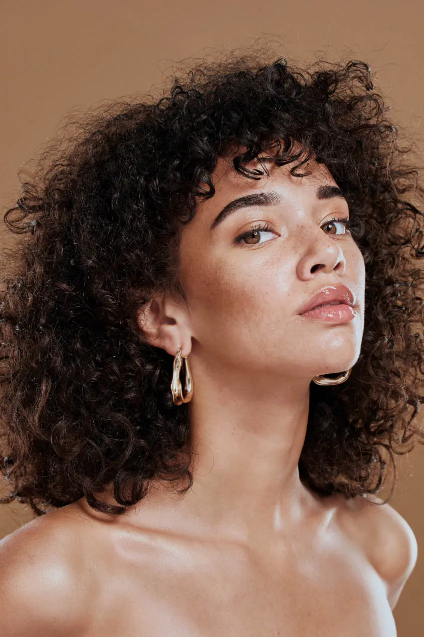A person with curly dark hair, styled impeccably as if by the best hair salon in Cary, NC, gazes confidently at the camera against a warm brown background. They are wearing large gold hoop earrings and have a subtle, natural makeup look. Their bare shoulders suggest an off-shoulder top.