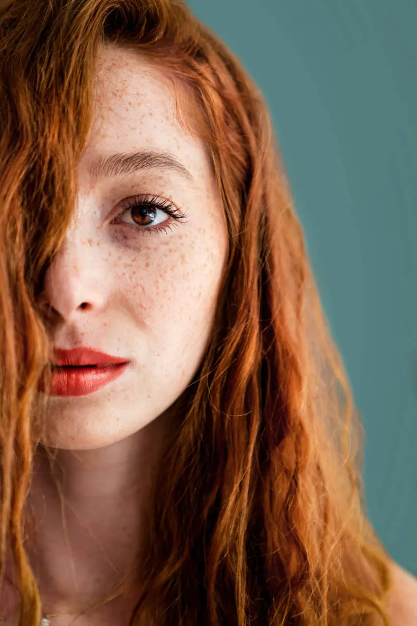 Close-up portrait of a woman with long, wavy red hair covering part of her face, against a teal background. She has freckles and is wearing red lipstick, with a calm and serious expression. If searching for hair salons in Cary NC to achieve this look, consider the best hair salons Cary NC offers.