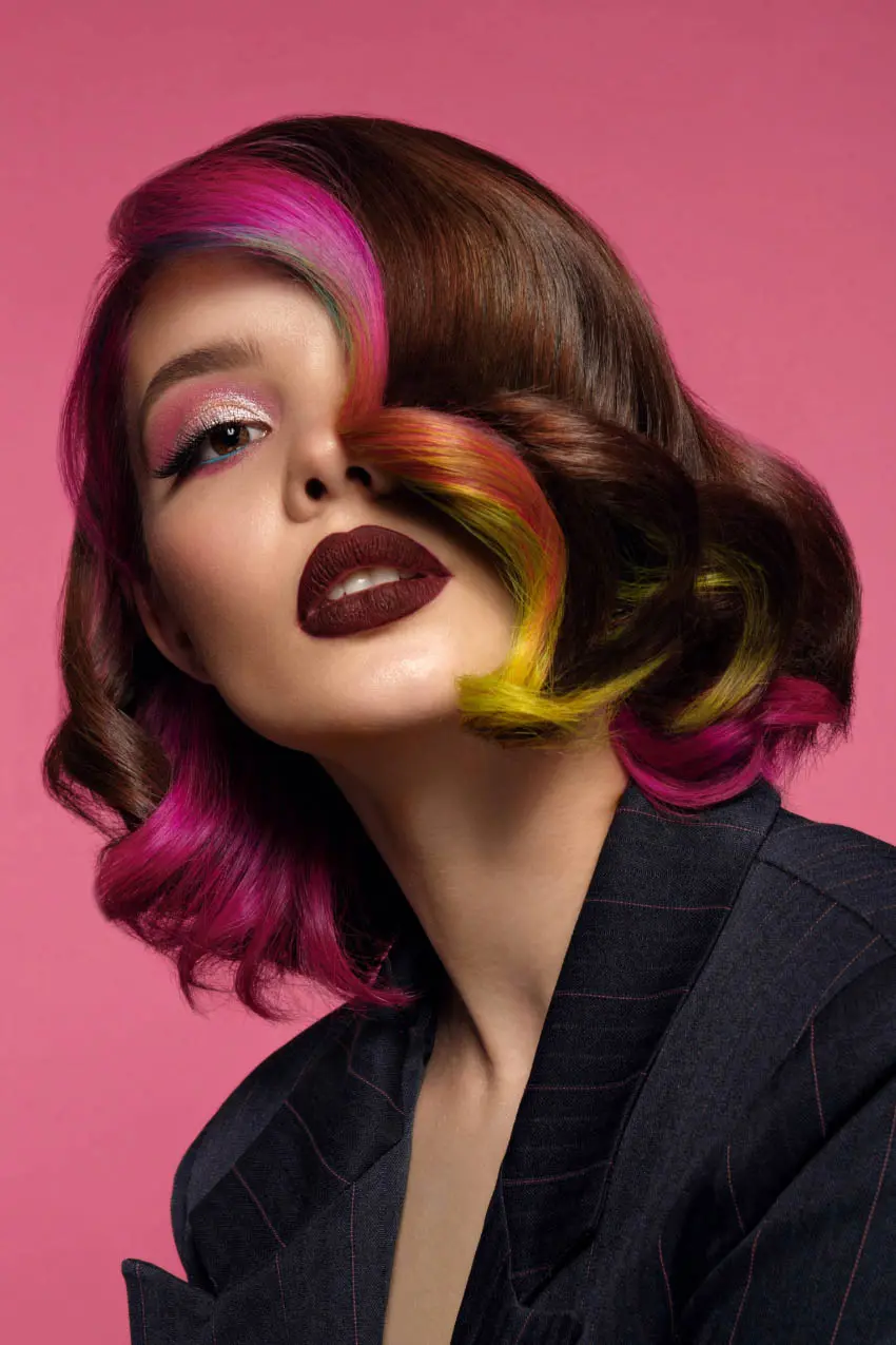 A person with shoulder-length wavy hair that has bright pink, yellow, and green streaks against a darker base poses against a pink background. They wear bold makeup, including dark lipstick and pink eye shadow, and a dark tailored jacket—perfect inspiration for your next visit to a hair salon near me.
