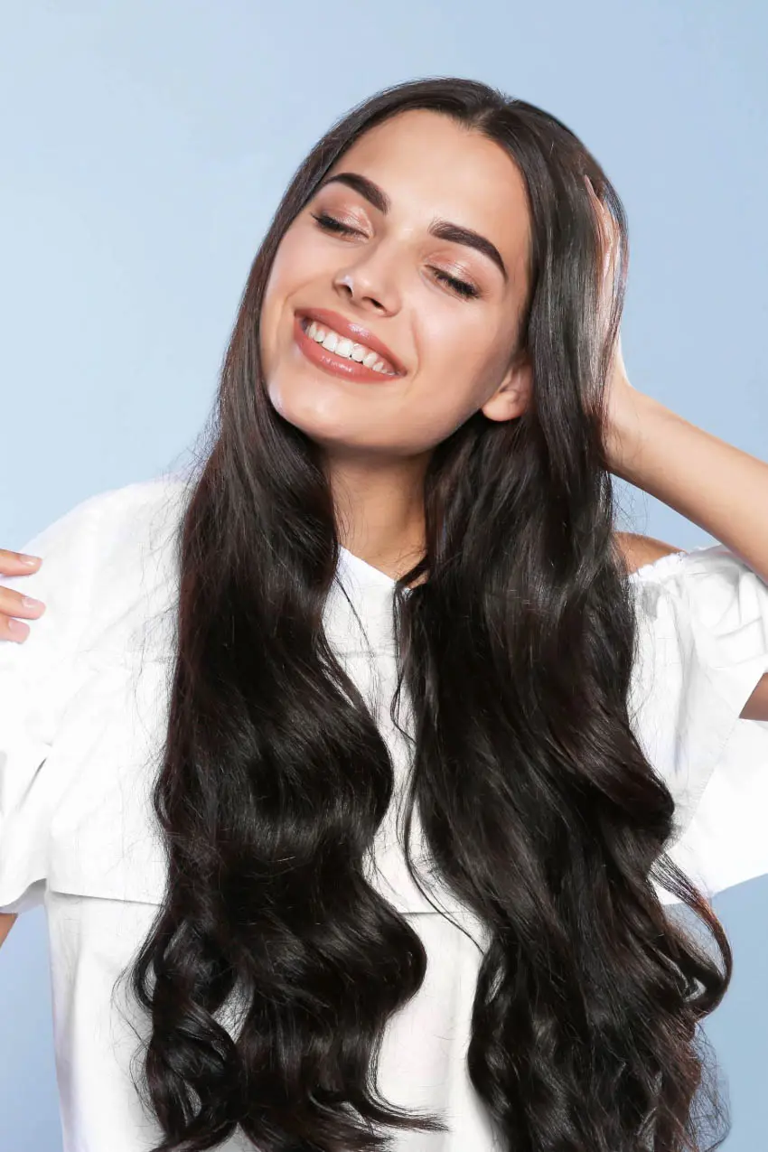 A woman with long, wavy, dark hair smiles with her eyes closed. She is wearing a white top and has one hand behind her head, with a light blue background behind her—just the kind of blissful vibe you'd find at Sunday Salon in Cary, NC.