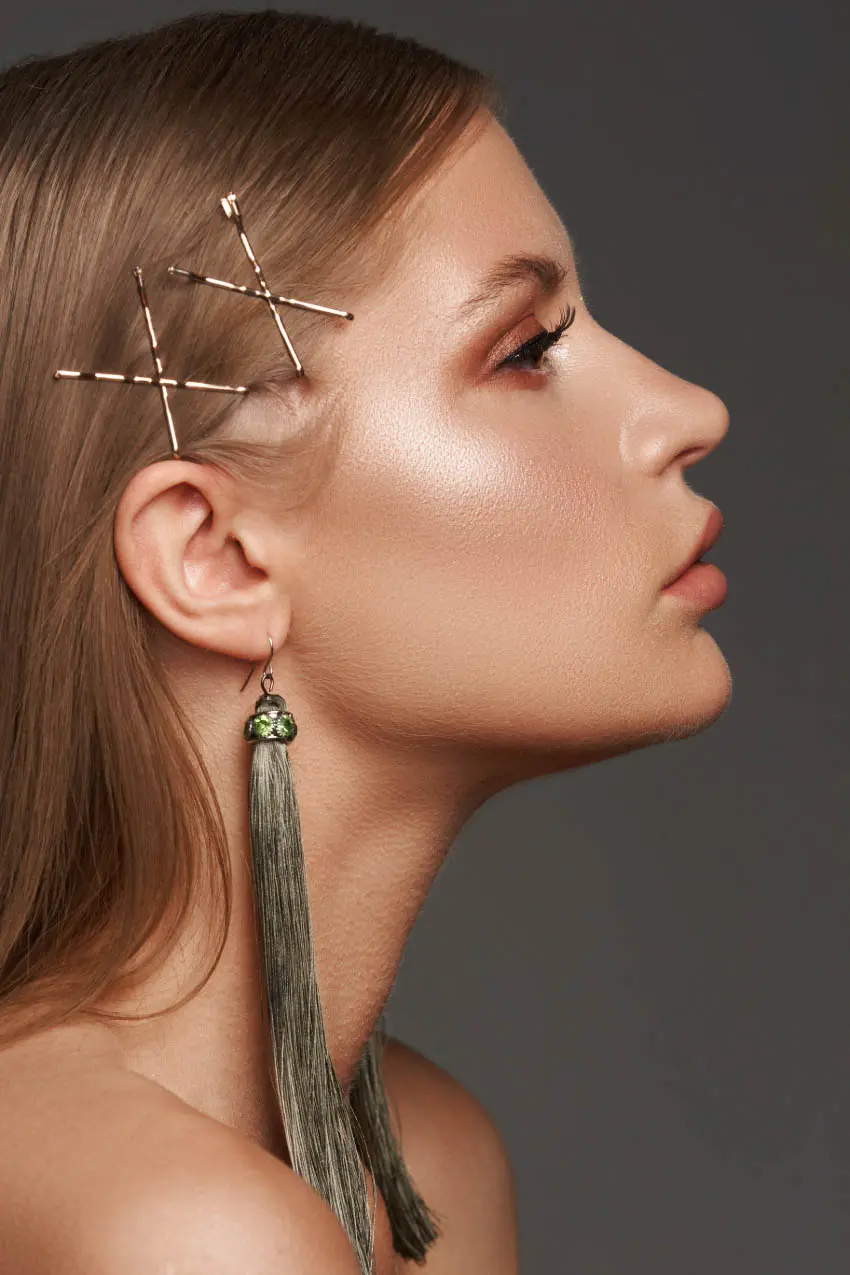 Profile view of a person with smooth skin and long hair, styled with crisscrossed bobby pins. They are wearing a long, green tassel earring and have shimmery makeup on their eyelids and cheeks. This chic look is perfect for visiting a hair salon near me. The background is a plain, dark gray.