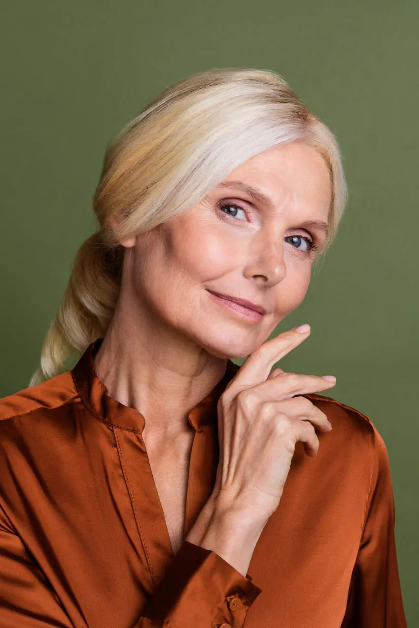 A mature woman with long, blonde hair tied back, wearing a silky, rust-colored blouse, is posing in front of a green background. She has a calm expression, with her hand gently resting on her chin, embodying the elegant vibe one might expect at a Sunday salon.