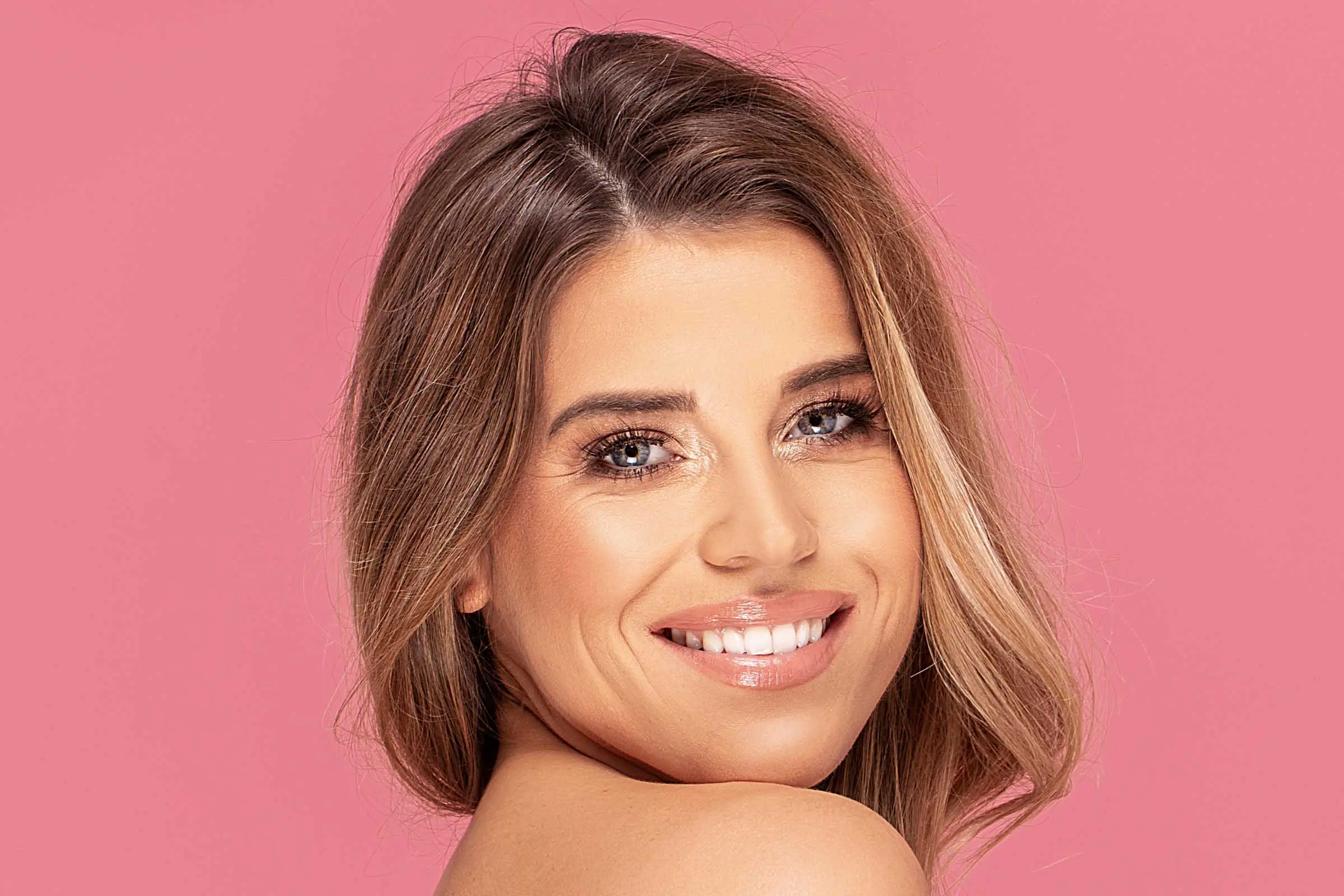A woman with light brown hair accented by a partial balayage smiles confidently at the camera. Her bright blue eyes and soft pink lipstick complement her natural makeup look. She is posed against a solid pink background, radiating charm in Cary, NC.