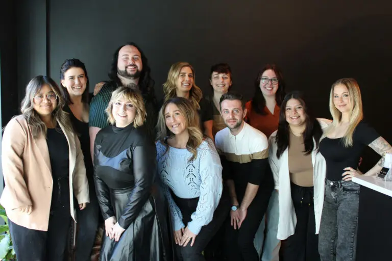 A group of eleven people posing for a photo against a dark background. They are standing and kneeling in two rows, all smiling. The group includes men and women with diverse clothing styles and hair colors, looking like they just stepped out of the best hair salon Raleigh, NC has to offer at Raleigh Iron Works.